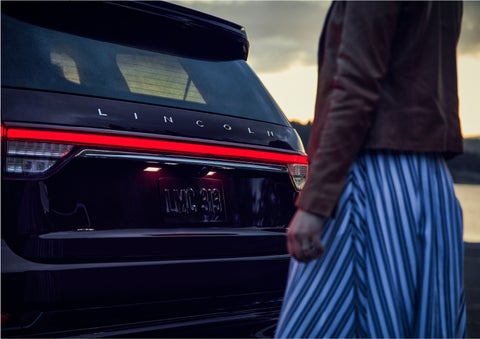 A person is shown near the rear of a 2023 Lincoln Aviator® SUV as the Lincoln Embrace illuminates the rear lights | Mike Reichenbach Lincoln in Florence SC
